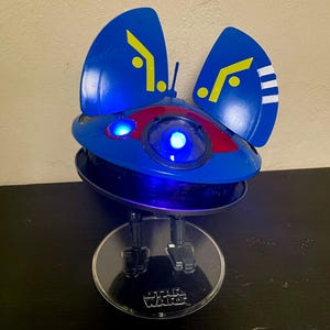 May include: A blue and red Star Wars toy spaceship with light-up features. The spaceship has two large blue wings with yellow markings and a clear plastic stand with the Star Wars logo.
