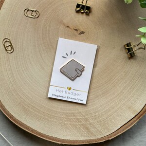 Könnte beinhalten: Eine weiße Emaille-Anstecknadel mit einem grauen Notizbuch-Design. Die Nadel befindet sich auf einer weißen Karte mit dem Text "Hei Budget Magnetic Enamel Pin".