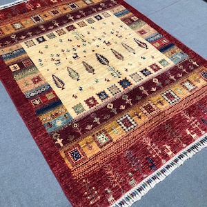 Puede incluir: Una alfombra rectangular, tejida a mano, con un campo central beige con motivos estilizados de hojas y árboles. La alfombra tiene un borde rojo oscuro con patrones geométricos y flecos. El diseño general es de estilo tribal tradicional.