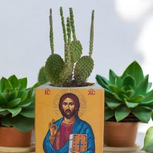 May include: A yellow wooden planter with a religious icon of Jesus Christ holding a book. The planter has the text "IC XC" on it. The planter is filled with a green cactus plant.