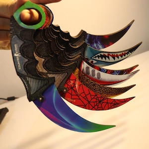 Op de afbeelding: Een set van vijf kleurrijke karambit-messen met verschillende ontwerpen. De messen hebben een zwarte handgreep en een gebogen lemmet. De ontwerpen omvatten een haai, een spinnenweb en een draak.