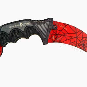 Op de afbeelding: Een zwart en rood karambit-mes met een spinnenwebpatroon op het lemmet. De handgreep is zwart en de woorden "COUNTER STRIKE" zijn aan de zijkant gedrukt. Het lemmet is gebogen en heeft een scherpe punt.