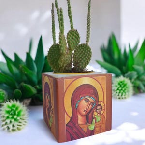 May include: A wooden planter with a religious icon of the Virgin Mary and baby Jesus. The planter is painted in shades of brown, red, yellow, and blue. A small cactus plant is growing in the planter.