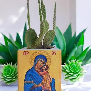 May include: A small, square, yellow planter with a painted image of the Virgin Mary and baby Jesus. The planter is filled with a green cactus plant. The planter has the letters "MP" and "OY" on the top corners.