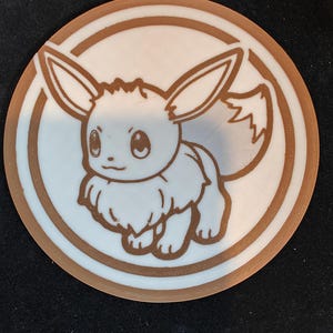 Op de afbeelding: Een ronde, bruin en witte coaster met een cartoon Eevee-personage. Het Eevee-ontwerp is bruin, met een witte achtergrond. De coaster heeft een bruine rand en ligt op een zwart oppervlak.