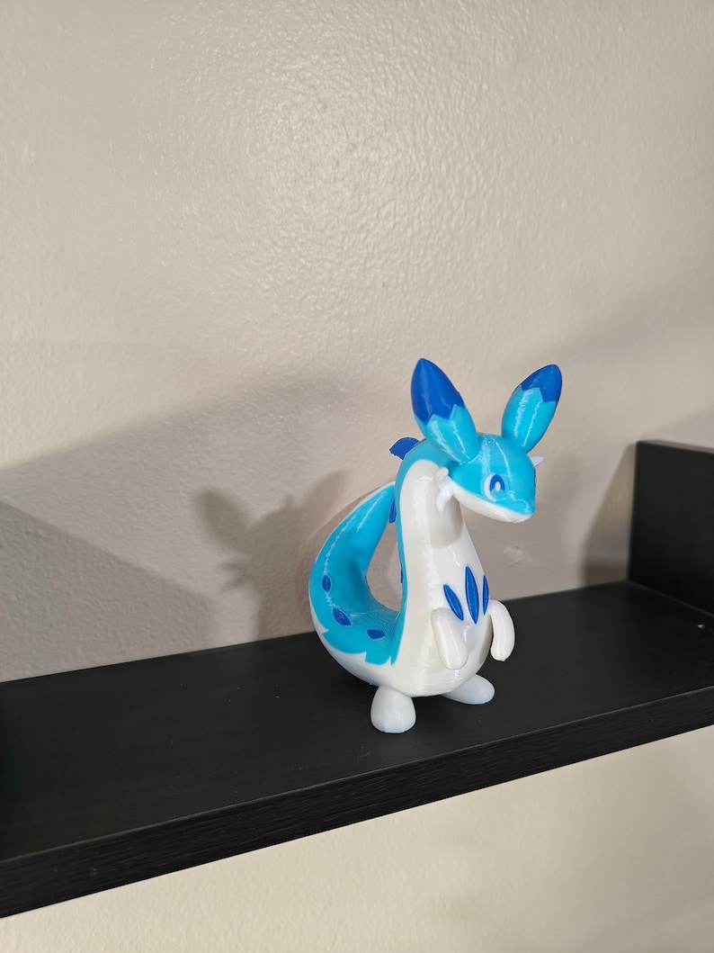 Puede incluir: Una figura de drag&oacute;n impresa en 3D de color blanco y azul, con orejas azules y cola enroscada.