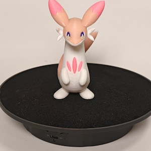May include: A white and pink 3D printed figurine of a fox-like Pokemon with purple eyes. The figurine is standing on a black rotating display base.