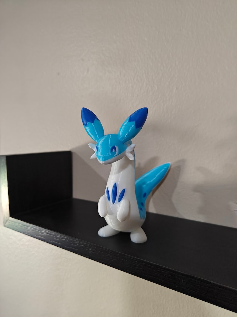 Puede incluir: Una figurita impresa en 3D de color azul y blanco de una criatura m&iacute;tica con cuello largo y cola. La criatura tiene orejas azules y un patr&oacute;n azul en su cuerpo.