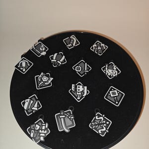 Könnte beinhalten: Eine Sammlung kleiner, quadratischer, schwarz-weißer Anhänger mit verschiedenen geometrischen und symbolischen Designs. Die Anhänger sind auf einer schwarzen, kreisförmigen Präsentationsfläche angeordnet, jeder mit einer kleinen Schlaufe. Das Design ist minimalistisch und grafisch.