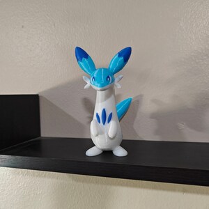 Puede incluir: Una figura de un personaje de Pokemon impresa en 3D en blanco y azul. El personaje tiene un cuello largo, un cuerpo blanco, orejas azules y una cola azul.