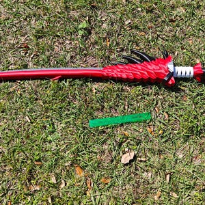 Puede incluir: Una espada de juguete roja y plateada con un diseño de dragón. La espada está tumbada sobre un lecho de hierba verde.