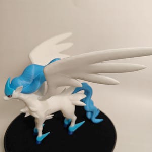 May include: A 3D-printed figurine of a mythical creature with white and blue coloring. The figure has large, white wings and stands on a black circular base. The creature has a horse-like body with a flowing blue mane and tail.