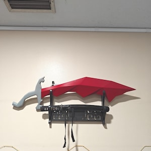 May include: A red, low-poly style sword with a grey handle mounted on a black display stand. Below the sword rests a black scabbard with a wrapped handle and hanging black cord, all positioned horizontally on a wall-mounted black wooden rack.