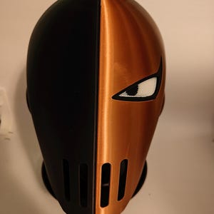 May include: A two-toned helmet, split vertically. One side is black, the other is metallic orange. The orange side features a white eye opening and vertical slits. This cosplay helmet is likely inspired by a comic book character.