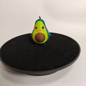 May include: A small green avocado figurine with a brown pit and a cute cartoon face. The figurine is standing on a black turntable.
