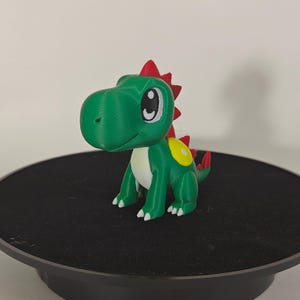 May include: A 3D-printed green dinosaur figurine with a red crest and yellow accents. The dinosaur has large, expressive eyes and white claws. The figurine is displayed on a black rotating platform.