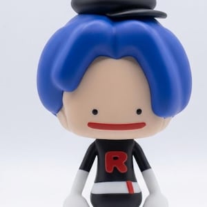May include: A stylized figurine with blue hair and a black cap. The figure wears a black shirt with a red "R" and black pants. White gloves and shoes complete the look. The figure has a simple, smiling face.