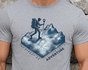 Könnte beinhalten: Ein graues T-Shirt mit einer blauen Grafik eines Wanderers mit Rucksack, der auf einem Berg mit Höhenlinien wandert. Die Grafik enthält auch das Wort "ADVENTURE".