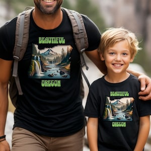 May include: A father and son wearing matching black t-shirts with a graphic of a waterfall and mountains in the shape of the state of Oregon. The text "Beautiful Oregon" is printed on the shirts.