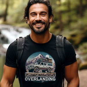 May include: Black crew neck t-shirt featuring a graphic of a 4x4 truck with camping gear, mountains, and the word "OVERLANDING" in white. The design is set against a blue and tan backdrop. The person is wearing a black backpack.
