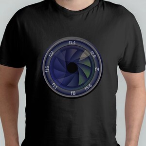 Könnte beinhalten: Ein schwarzes T-Shirt mit einem Aufdruck eines Kameraobjektiv-Blendenrings. Die Blende ist ein Kreis mit einem silbernen Rand und einer blauen und grünen Iris. Die Zahlen f1.4, f2.8, f4, f5.6, f8, f11, f16 und f22 sind um die Blende herum gedruckt.