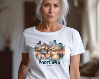Könnte beinhalten: Ein weißes T-Shirt mit einer farbenfrohen Illustration der Skyline von Portland, Oregon, die sich im Wasser spiegelt. Das Wort "PORTLAND" ist in fetter, dunkler Schrift gedruckt, darunter "OREGON". Das T-Shirt wird von einer Person mit silbernem Haar getragen.