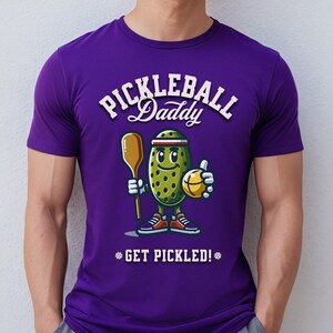 Könnte beinhalten: Ein lila T-Shirt mit dem Text "PICKLEBALL Daddy" in weißer Schrift über einer Cartoon-Gurkenfigur, die einen Schläger und einen Ball hält. Der Satz "GET PICKLED!" steht darunter.
