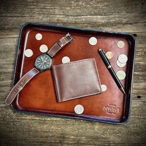 May include: A rectangular brown leather valet tray with dark edges, featuring the embossed text "MADE BY HERITAGE LEATHER". Inside the tray are a brown leather wallet, a black wristwatch with a brown leather strap, a black pen, and several silver coins.