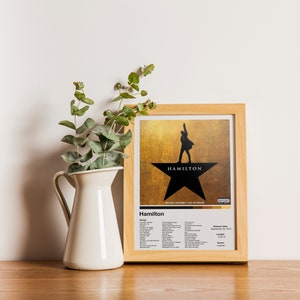 May include: A framed print of the Hamilton musical poster. The poster features a silhouette of Alexander Hamilton standing on a star with the word "Hamilton" written on it. The poster is in a wooden frame and is sitting on a wooden table.