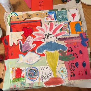 May include: A collage of colorful children's drawings on paper, featuring a central figure with a pink spiky head, blue torso, and yellow tail. Surrounding elements include a red heart, a purple handprint, a fish, and various small sketches.