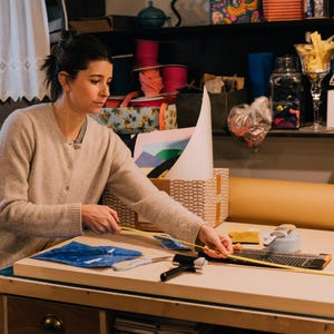 May include: A beige button-down cardigan worn over a gray shirt, with a yellow measuring tape extended across a large rectangular wooden board, alongside a black brayer, scissors, and a paper trimmer on a workspace surface.