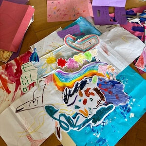 May include: A collection of children's artwork featuring various drawings and paintings on paper. The pieces include colorful rainbows, flowers, a sun, and abstract shapes in markers and paint. Some papers have handwritten text and cut-out shapes.
