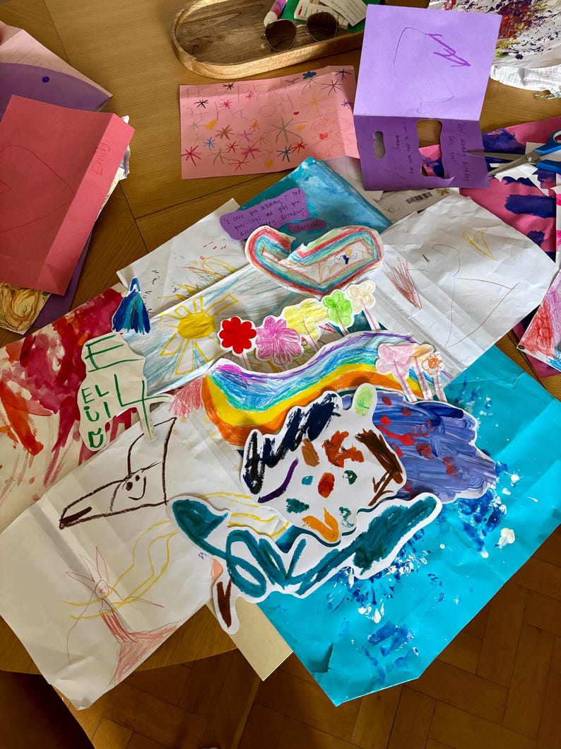 May include: A collection of children's artwork featuring various drawings and paintings on paper. The pieces include colorful rainbows, flowers, a sun, and abstract shapes in markers and paint. Some papers have handwritten text and cut-out shapes.