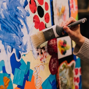 May include: A paint brush with a grey handle and silver ferrule applying blue paint onto a large collage board. The board features various colorful paper cutouts, including red circles, orange shapes, and blue scribbles on a white background.