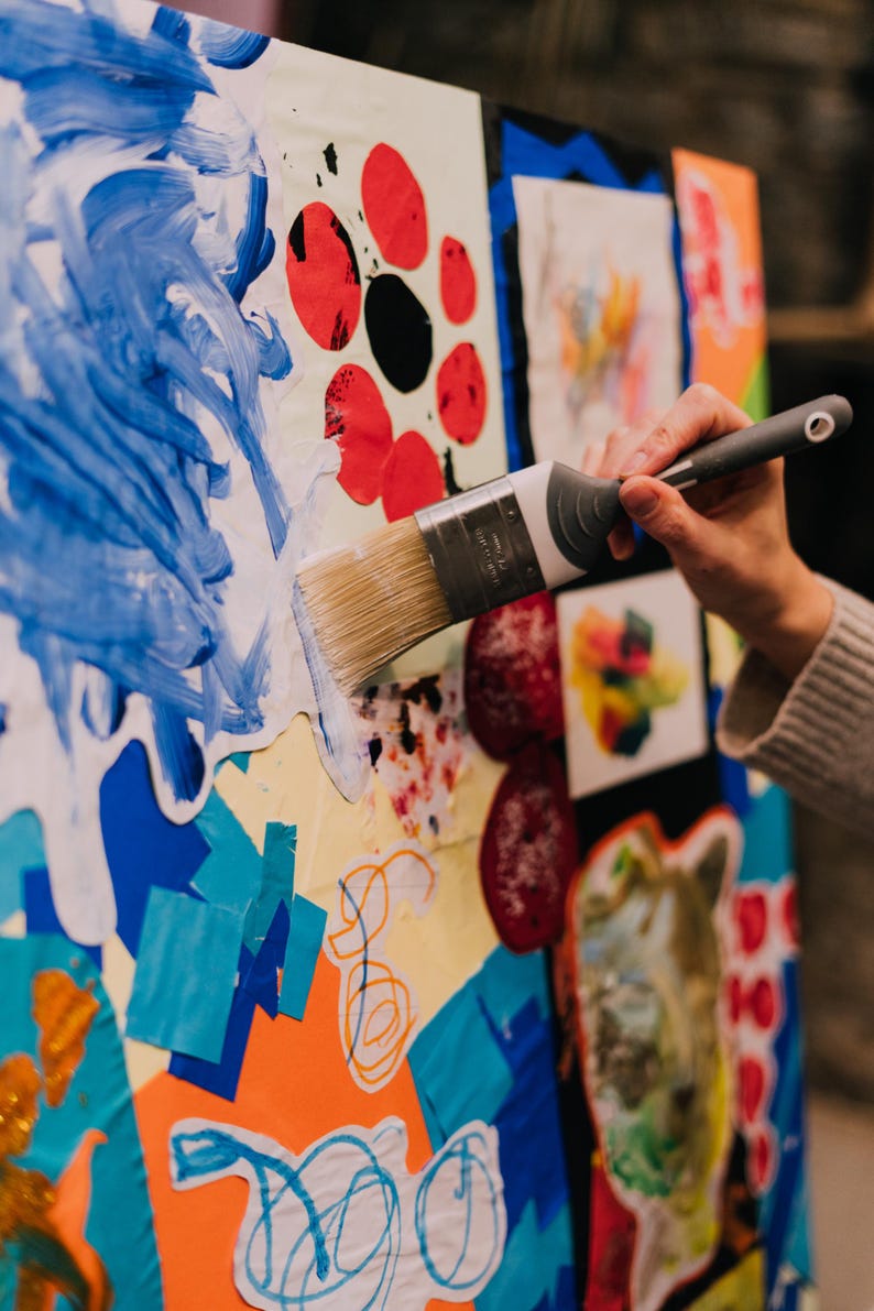 May include: A paint brush with a grey handle and silver ferrule applying blue paint onto a large collage board. The board features various colorful paper cutouts, including red circles, orange shapes, and blue scribbles on a white background.