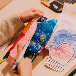 May include: A pair of light gray scissors cutting through a sheet of multicolored tissue paper with blue, orange, and pink sections. Nearby lies a white paper cutout of a mitten featuring colorful scribbles in orange, pink, and blue ink.