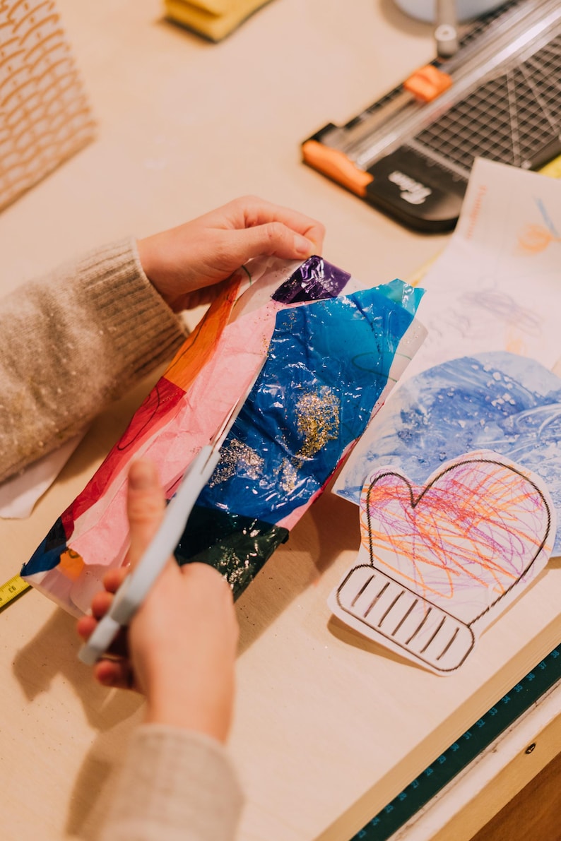 May include: A pair of light gray scissors cutting through a sheet of multicolored tissue paper with blue, orange, and pink sections. Nearby lies a white paper cutout of a mitten featuring colorful scribbles in orange, pink, and blue ink.
