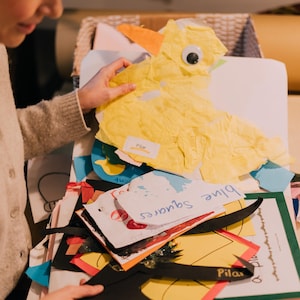 May include: A collection of colorful paper craft projects including a yellow tissue paper duck with a googly eye, a white paper cutout labeled blue squares, and various other shapes in black, red, and yellow with the name Pilar written on them.