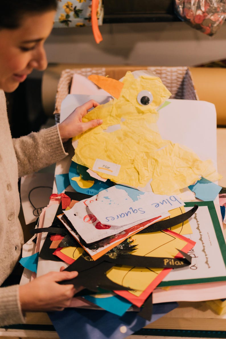 May include: A collection of colorful paper craft projects including a yellow tissue paper duck with a googly eye, a white paper cutout labeled blue squares, and various other shapes in black, red, and yellow with the name Pilar written on them.