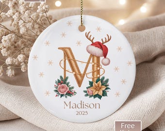 May include: White ceramic Christmas ornament with a gold letter "M" decorated with a reindeer antler Santa hat and floral accents. The name "Madison" and the year "2025" are printed below the letter. The ornament hangs from a gold string.
