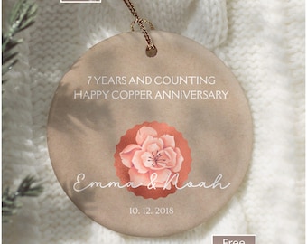 May include: A round, light brown ornament with the text "7 YEARS AND COUNTING HAPPY COPPER ANNIVERSARY" and the names "Emma & Noah" with the date "10. 12. 2018". A copper-colored flower is in the center. A gold string is attached.