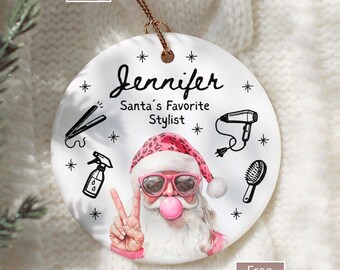 May include: A white ceramic Christmas ornament with a Santa Claus design. Santa is wearing pink sunglasses and a pink Santa hat, blowing a bubblegum bubble. The ornament says "Jennifer Santa's Favorite Stylist."
