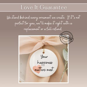 May include: A white, round ornament with the text "Your happiness matters most" and a pink heart. The ornament hangs from a gold string and a peach-colored ribbon. A "Satisfaction Guarantee" seal is visible.