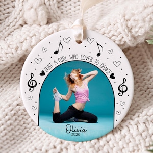 May include: A round white ceramic ornament featuring a central photograph of a dancer in a pink tank top and black pants. The text reads "JUST A GIRL WHO LOVES TO DANCE", "Olivia", and "2026". Black musical notes and hearts decorate the border.