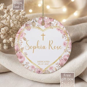 May include: A round, white ceramic ornament with a floral design and gold accents. The ornament features the name "Sophia Rose" in gold script, a cross, and the date "05.07.2027". Pink roses and greenery surround the heart-shaped center.
