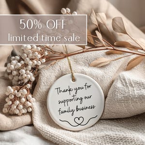 May include: A white ceramic ornament with a gold string, featuring the text "Thank you for supporting our family business" in black script. The ornament is set against a soft, neutral-toned background with dried foliage and the text "50% OFF limited time sale".