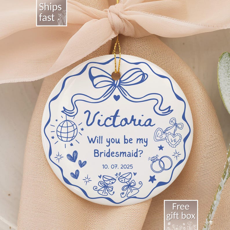 May include: A white ceramic ornament with blue decorative elements and text. The ornament says "Victoria, Will you be my Bridesmaid?" with the date 10. 07. 2025. The ornament is tied with a pink ribbon.
