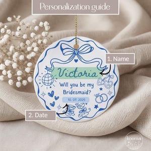 May include: A white ceramic ornament with a blue ribbon design and the text "Victoria, Will you be my Bridesmaid?" and the date "10. 07. 2025". The ornament has a gold string for hanging and is decorated with hearts, stars, and a disco ball.