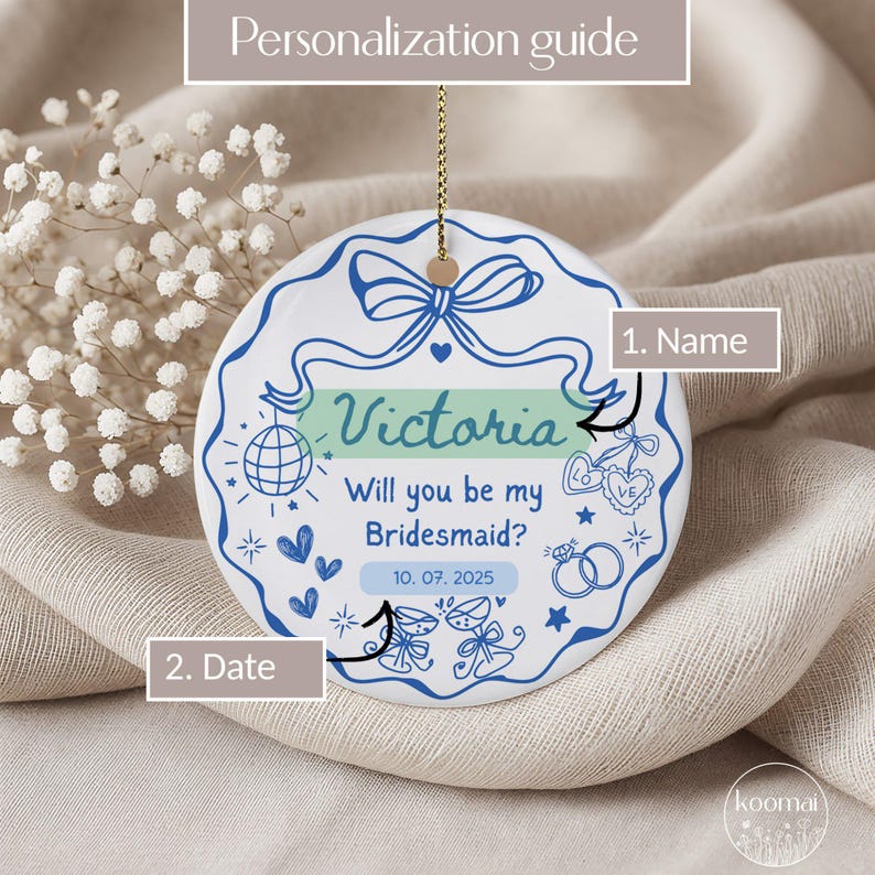 May include: A white ceramic ornament with a blue ribbon design and the text "Victoria, Will you be my Bridesmaid?" and the date "10. 07. 2025". The ornament has a gold string for hanging and is decorated with hearts, stars, and a disco ball.