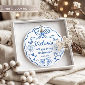 May include: A white ceramic ornament with blue text and designs, presented in a white gift box. The ornament features the name "Victoria" and the question "Will you be my Bridesmaid?" along with a date and decorative elements.